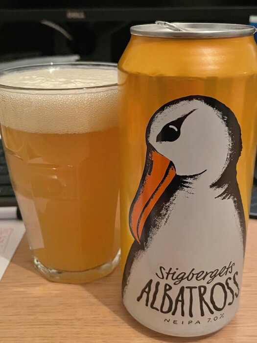 En ölburk av märket Stigbergets Albatross NEIPA 7.0% bredvid ett glas med skummande öl på ett träbord.