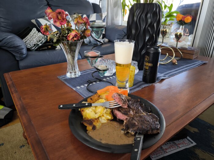 Tallrik med entrecote, potatisgratäng, grönpepparsås och paprika på ett soffbord, med ett glas öl och blommor i bakgrunden.