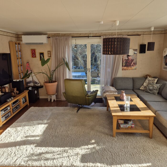 Vardagsrum med grön fåtölj, soffa, stort fönster, tv och inomhusväxt. Luftvärmepump syns ovanför bokhylla på väggen.