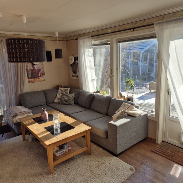 Vardagsrum med grå hörnsoffa, träbord och stora fönster som släpper in ljus. Filta och kuddar på soffan, samt växter vid fönstret.