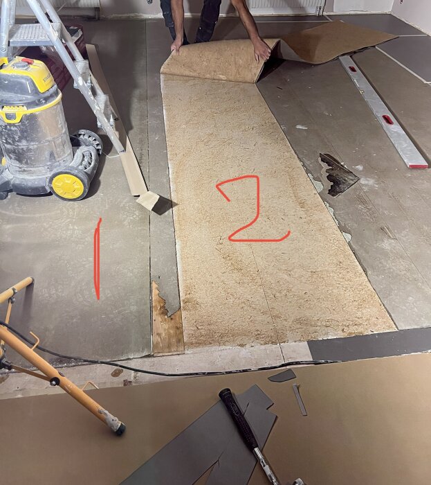 Person removes vinyl and foam sheet, revealing uneven plywood floor underneath; tools, ladder, vacuum visible nearby.