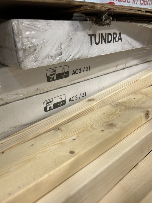 Förpackning märkta "Tundra" och "AC 3/31" med symboler för slitageklass, ovanpå träplankor.