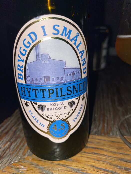 Flaska med etiketten "Bryggd i Småland, Hyttpilsner" från Kosta Bryggeri, placerad på ett träbord.