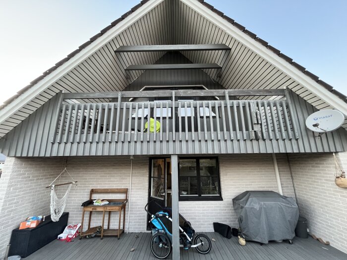 Gavelbalkong på ett 70-talshus med gråribbad balkongfront ovanför stensatt uteplats med hammock och grill.