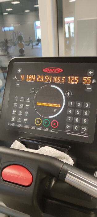 Närbild av en motionsmaskin display som visar kalorier, tid, avstånd, effekt och hastighet. En nödstopp-knapp och handduk syns också.