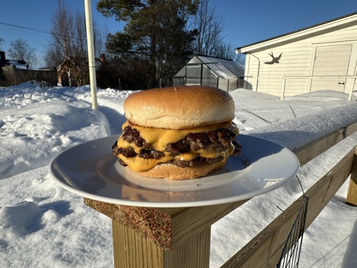 Hamburgare med ost på tallrik utomhus i snöigt landskap, soligt väder och trästaket i bakgrunden.