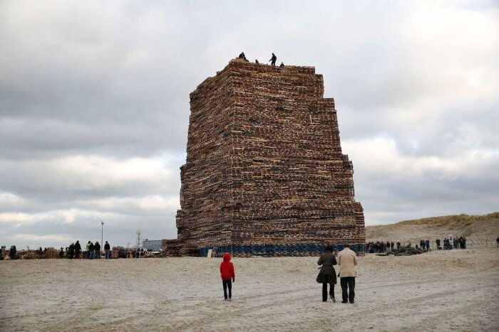 Enorma staplade träpallar bildar ett torn, med människor klättrandes längst upp, på en strandplats med molnig himmel.