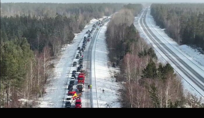 Bilkö och utryckningsfordon på snöig väg, medan andra körfältet är tomt. Skogsområde på båda sidor om vägen.