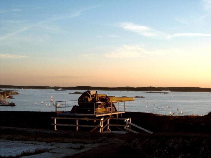 Kanonen vid Oscar II fortet med utsikt över havet i solnedgången, omgiven av segelbåtar.