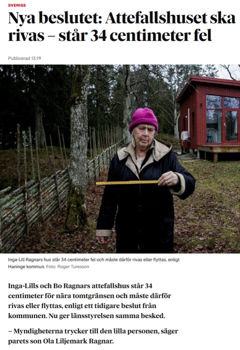 En person med rosa mössa håller en mätsticka framför ett rött attefallshus i bakgrunden, omgiven av träd och gärdsgård.