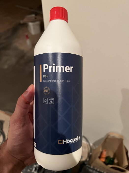 Flaska med Höganäs Primer FB5, koncentrerad primer 1 kg, hålls i en hand. Bakgrund visar byggmaterial.