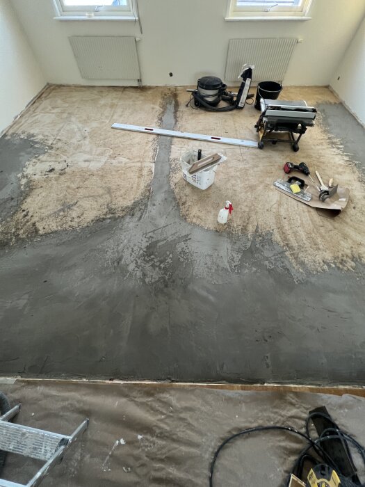 Rum med trägolv täckt med Byggspackel, verktyg, dammsugare och strykjärn står i rummet; nivåutjämningsprocess pågår.