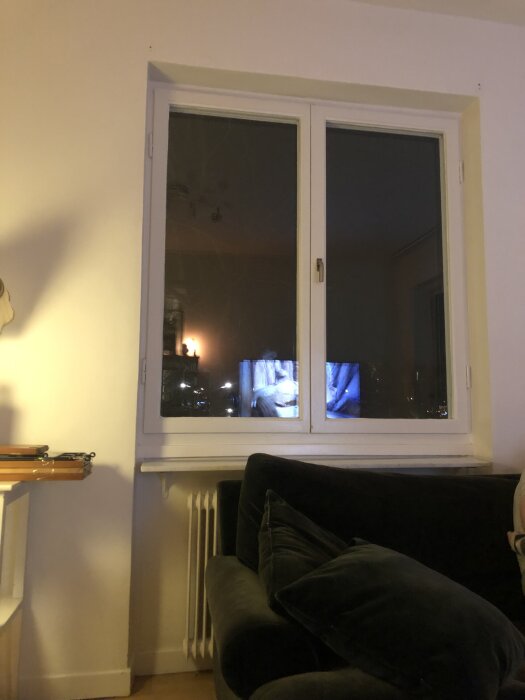 Vitt fönster med vit karm och vägg, svart soffa nedanför, samt en synlig fönsterbräda som reflekterar en inomhusbild på skärmen.