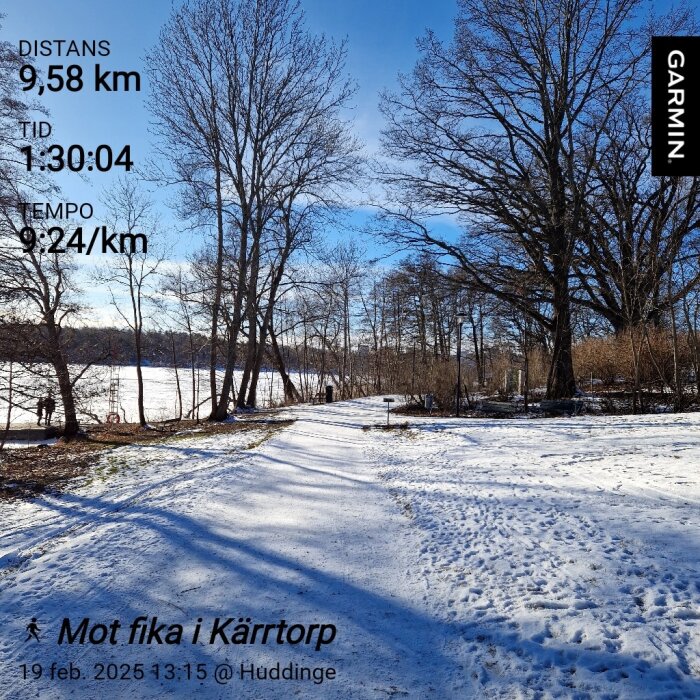 Snöig väg genom skog med synliga fotavtryck. Träd utan löv på båda sidor, blå himmel i bakgrunden. Text om distans, tid och tempo från Garmin.