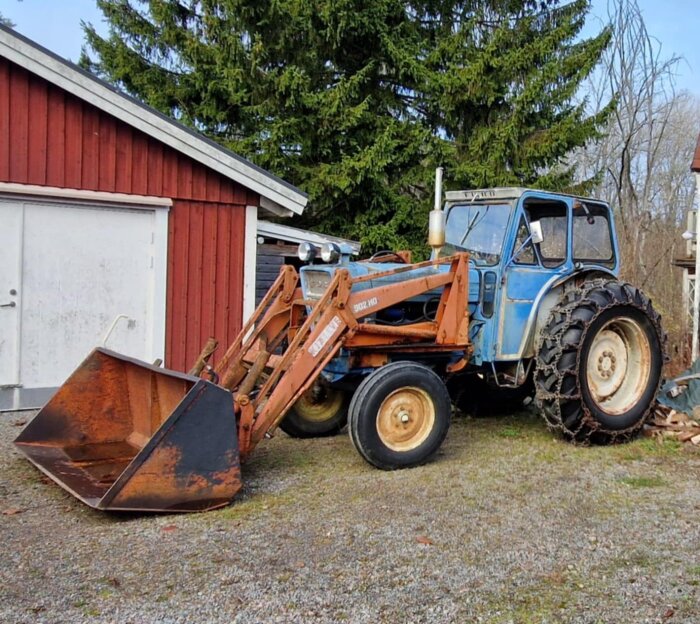 En blå Ford 5000 traktor från 1969 med frontlastare och snökedjor, parkerad vid en röd byggnad.