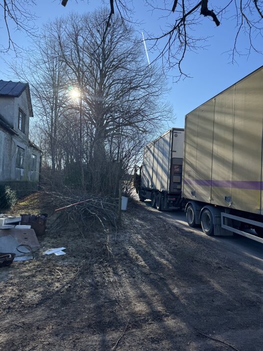 Lastbil utan gavellift levererar byggställning vid ett hus. Solig dag med träd i bakgrunden och mark belamrad med föremål.