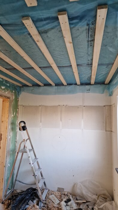 Stege står i ett rum med nylagd isolering och byggfolie i taket, väggar klara för spackling och målning, material på golvet.