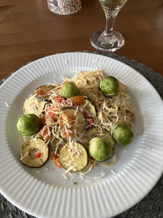Pasta med zucchini, tomat, chili, vitlök, citron och brysselkål, serverad på en vit tallrik, toppad med riven ost, med ett glas vin i bakgrunden.