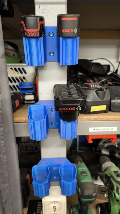 3D-printade blå hållare för Bosch 12V-batterier monterade på en hylla i verkstadsmiljö.