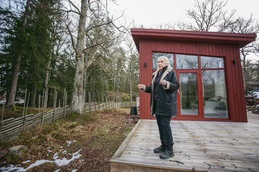 En man står på en altan vid ett rött hus, med ett staket och skog i bakgrunden.