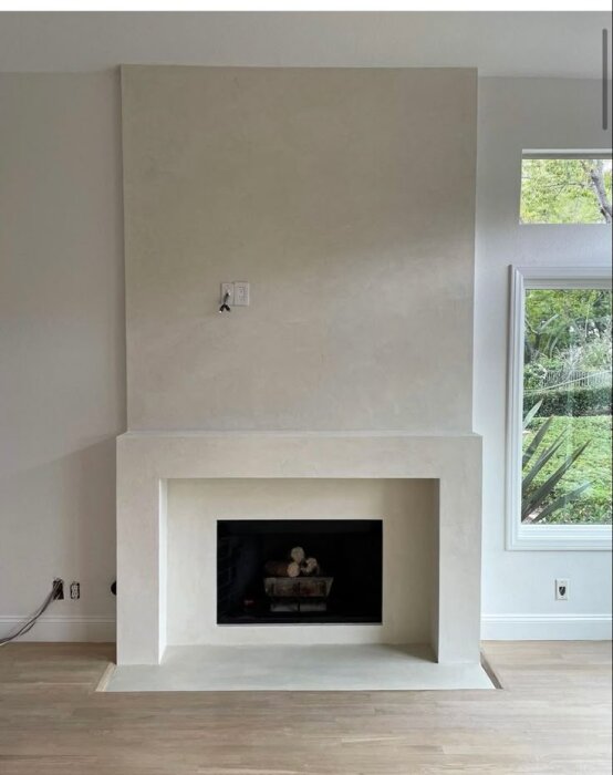 Slät putsad vägg med enfärgad yta och en öppen spis i modern design, placerad framför ett fönster med utsikt över trädgård.