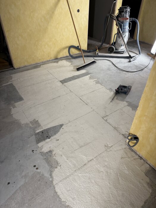 Rum med renoveringsarbete, dammsugare och borste på ett delvist avskrapat golv.