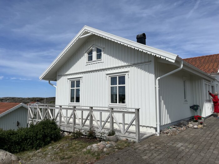 Vitt trähus med vita fönster av trä och vitlackad al-plåt, beläget nära havet på västkusten. En person i röd jacka står vid väggen.