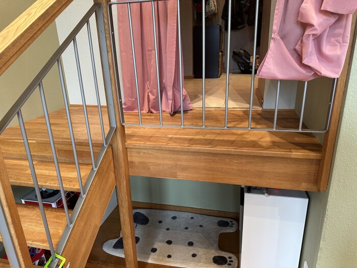 Puiset portaat, joiden yläosassa on pieni alusta. Näkyvissä metallikaiteet ja vaaleanpunainen verho. Alusta irtoamassa seinästä.