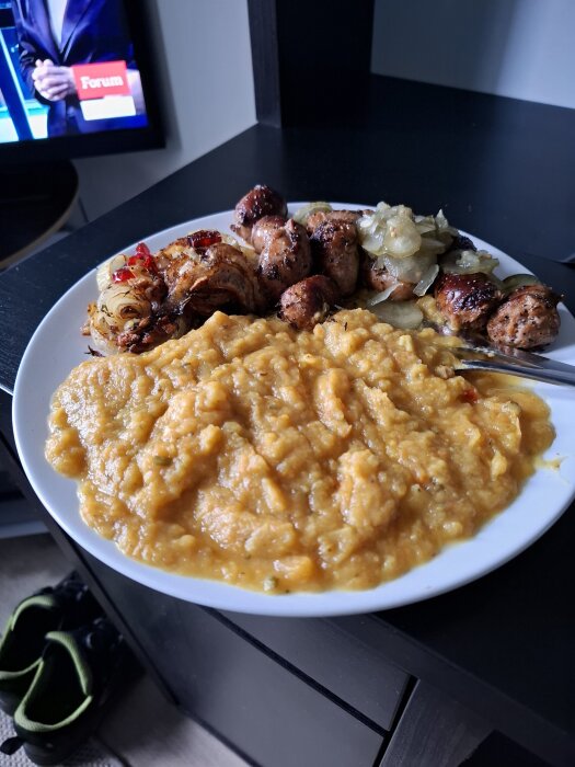 Tallrik med rotmos, salsiccia, stekt lök och inlagd gurka på ett bord. En TV är på i bakgrunden.
