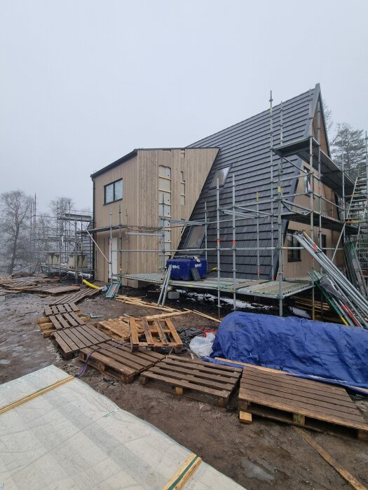 Byggarbetsplats med hus under konstruktion och ställning delvis nedmonterad. Pall och blå presenning syns på marken framför huset.