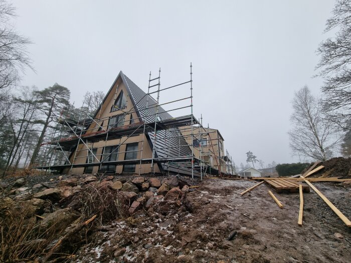 En villa med pågående rivning av byggställning. Bredvid huset syns staplade träbrädor och en del av en grusig mark med stenar.