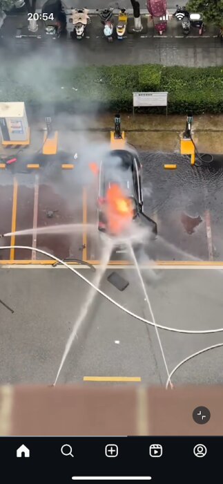Bil brinner på parkeringsplats, omgiven av fyra vattenstrålar som försöker släcka elden.