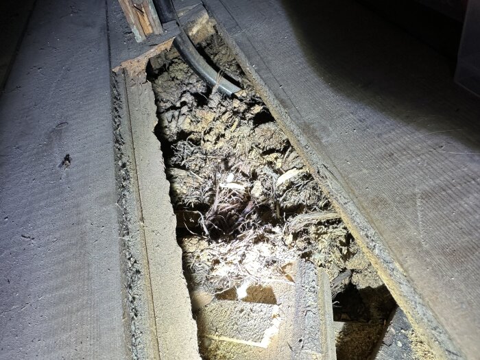 Bilden visar en snedställd träkonstruktion med sågspån och takpapp. Det finns en öppning i träskivorna som exponerar materialet under.