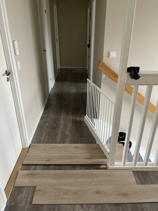 Laminatgolv läggs på tvären i en hall, plankor förberedda för installation, med trappräcke och dörrar synliga i bakgrunden.