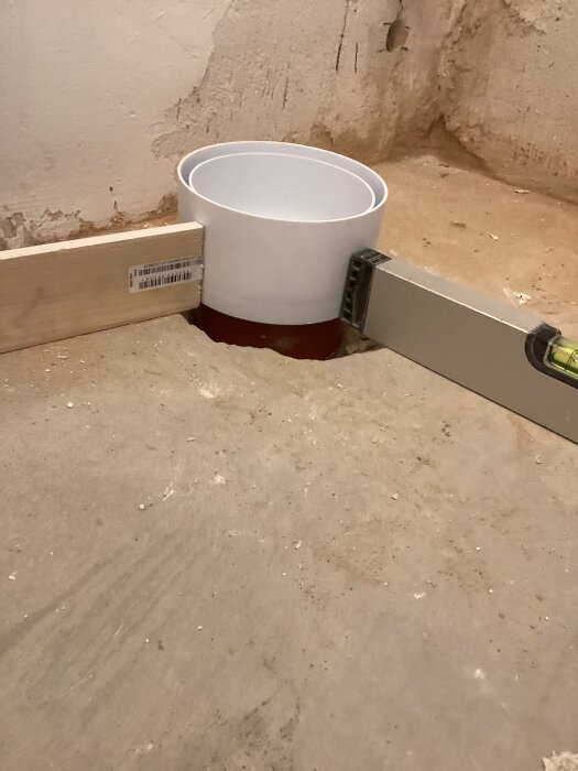 Faluplast WC-anslutning installerad i ett betonggolv, omgiven av en vattenpass och en träbit mot en slät vägg.