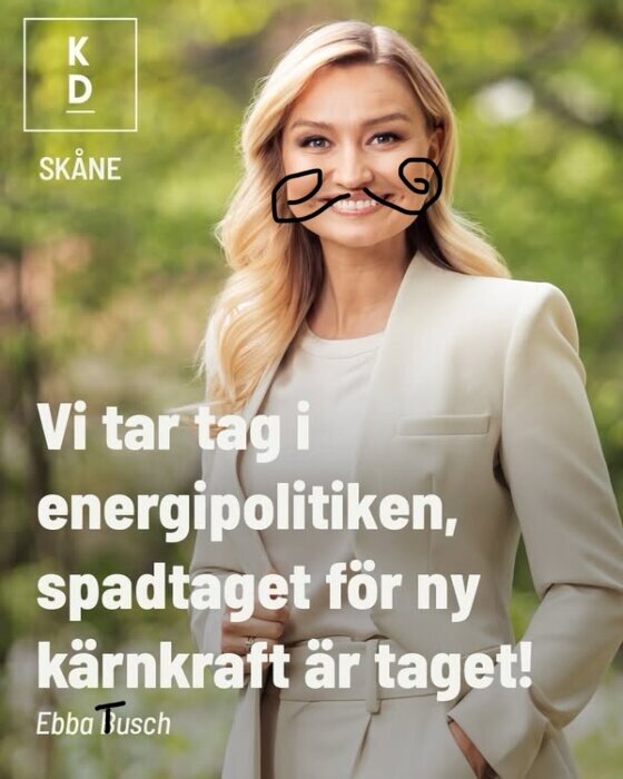 En kvinna i ljus kostym med tecknad mustasch står framför en suddig grön bakgrund. Text om energipolitik och kärnkraft syns på bilden.