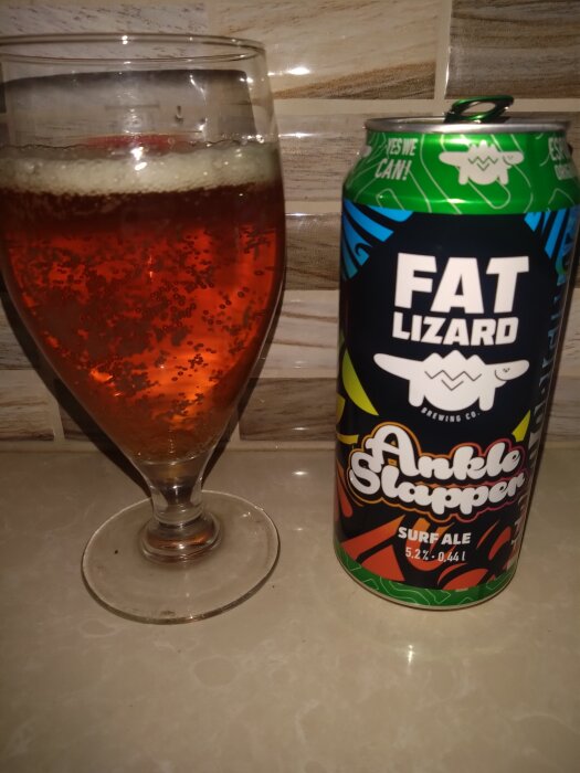 Ett glas med bärnstensfärgad öl bredvid en burk med etiketten 'Fat Lizard Ankle Slapper Surf Ale' på ett beige bord.