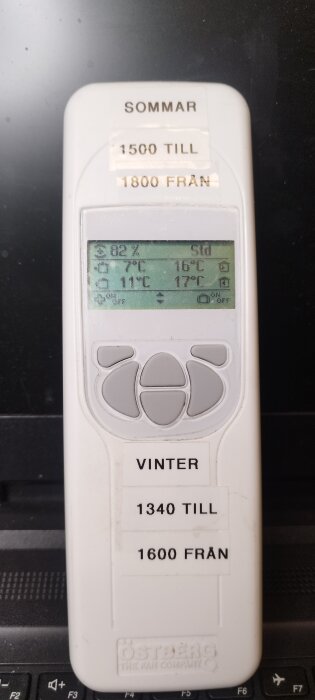 Vit fjärrkontroll med digital display, visar temperaturer och inställningar. Etiketter: "SOMMAR" och "VINTER" med tidsangivelser för till- och frånkoppling.