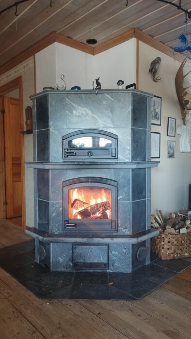 Vedeldad kamin i en hörna av rummet, med eld som brinner bakom glasluckan. Vedkorg med ved bredvid kaminen.