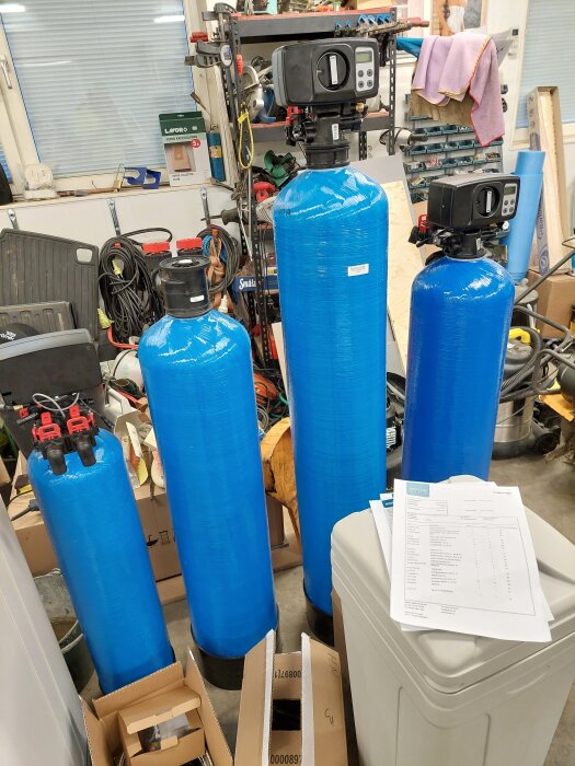 Fyra blå filtertankar med automatiska backspolningssystem i ett garage; dokument ligger ovanpå en vit behållare närmast kameran.