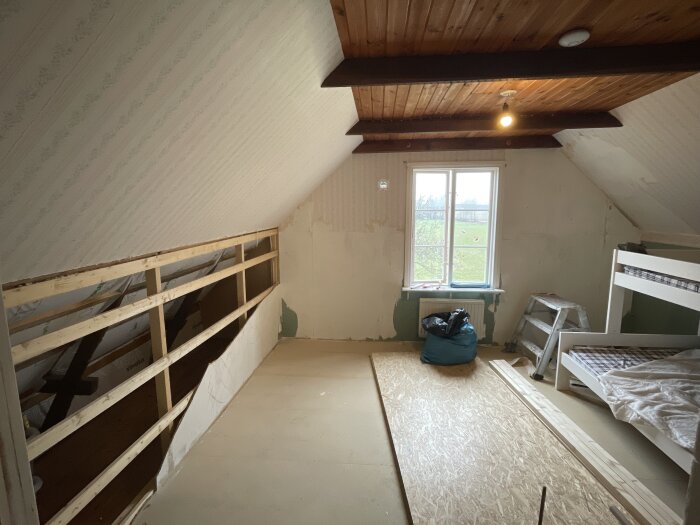Renoverat sovrum med öppnad kattvind, synliga trästolpar längs med taket, fönster, stege, säng och byggmaterial på golvet.