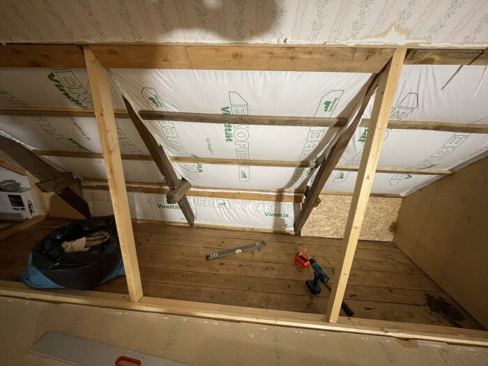 Öppet utrymme med träbalkar och takbjälkar, inspektionsverktyg och isolering synliga. En påse och verktyg ligger på golvet.