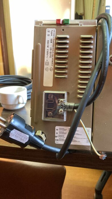 Bakpanelen på en elektrisk enhet med märkning och anslutna kablar, inklusive en strömkabel med kontakt.