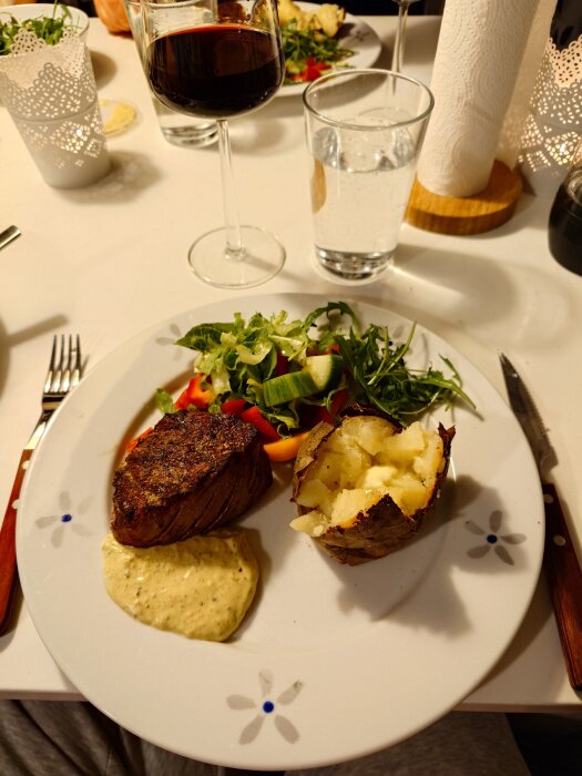 Tallrik med grillad rostas, bakad potatis, grönsaker och bearnaisesås. Glas med vin och vatten bredvid.
