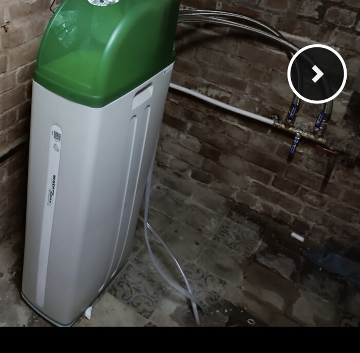 Vattenavhärdare installerad i källare med stenväggar och golv, ansluten med rör och slangar.