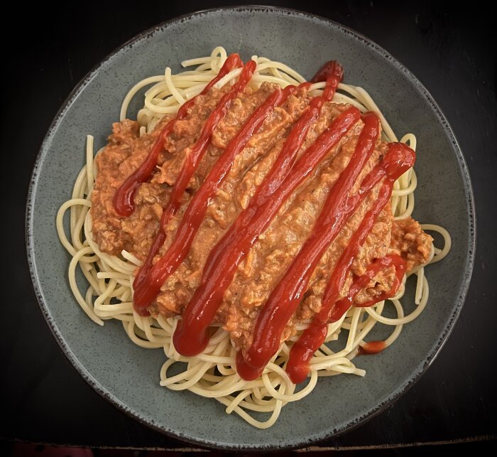 Spaghetti med köttfärssås toppad med ketchup på en grå tallrik.