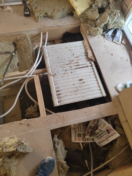 Öppet golv med en borttagen sektion som exponerar isolering, rör och en liggande gammal radiator. Tidningar och verktyg syns också.