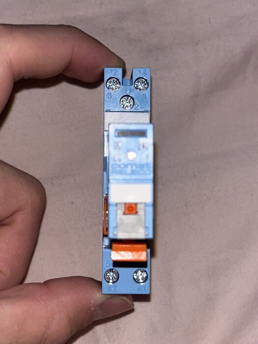 Blått relä för 24/230V med anslutningar märkta 11, 12, 14 för L och N, samt A1 och A2 längst ned, hålls mellan fingrar.