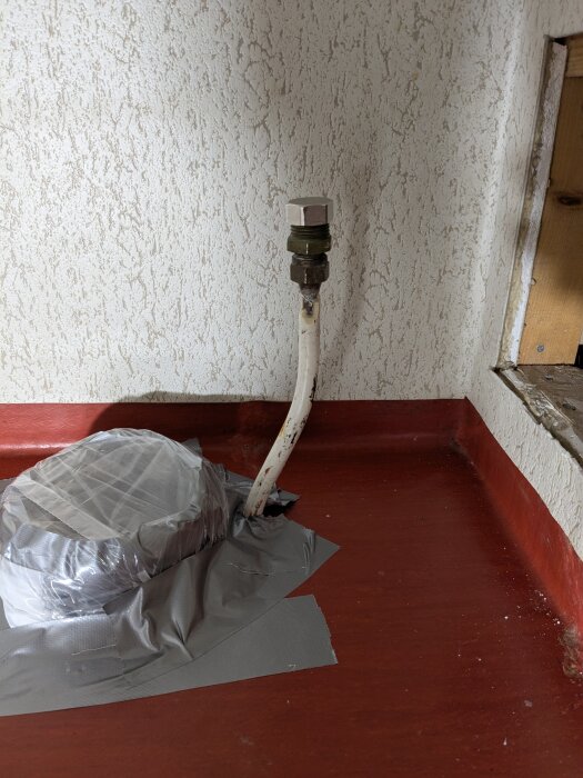 Rör i källare med silvertejp runt anslutning, mot vit strukturerad vägg och rött golv. Rör planeras att flyttas och böjas för toalettinstallation.