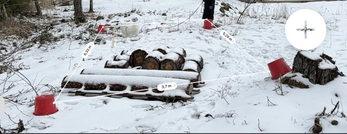 Snötäckt mark med uppmärkta avstånd för växthusbygge. Röda hinkar och vedträn markerar område, 3,7 m gånger 6,33 m. Trädstubbe syns närmast.